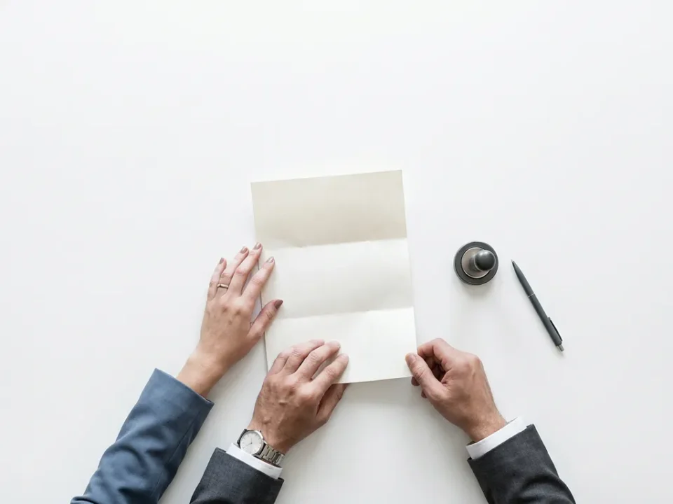 Twee sets handen die een formeel arbeidscontract vasthouden boven een wit bureau, met een bedrijfszegel en pen ernaast.