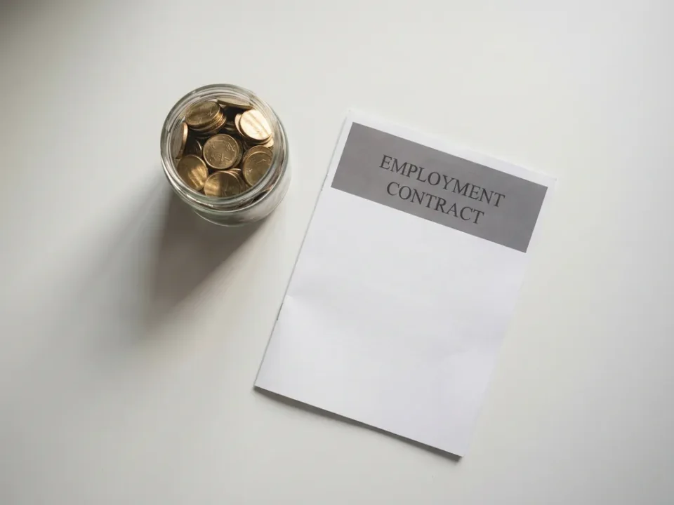Glazen pot gevuld met gouden munten naast een gevouwen arbeidscontract op wit bureau, zacht natuurlijk zijlicht.